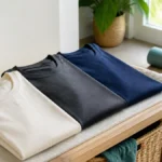 3 Everyday T-Shirts for an Active Lifestyle (2026) Three unbranded ivory, charcoal and navy T-shirts beside a water bottle, walking shoes and exercise mat.